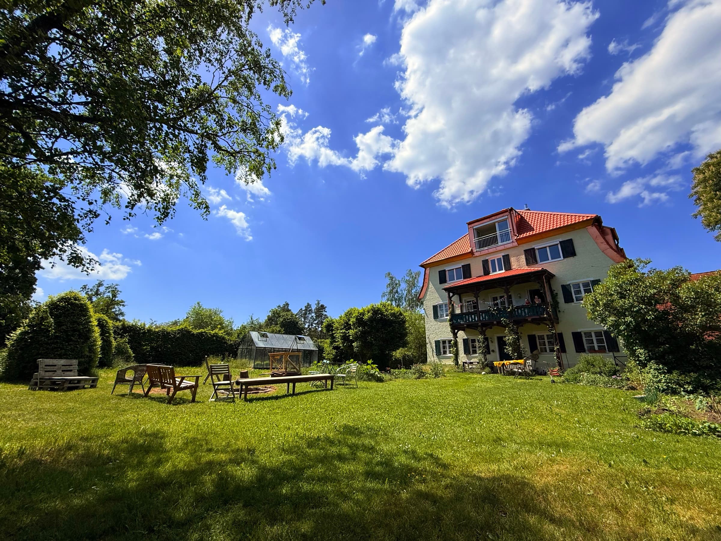 Langer Holztisch im Garten des Zehnerhofs mit Bänken, daneben ein Gewächshaus, im Hintergrund das Hofgebäude und ein weiter Himmel mit Wolken.