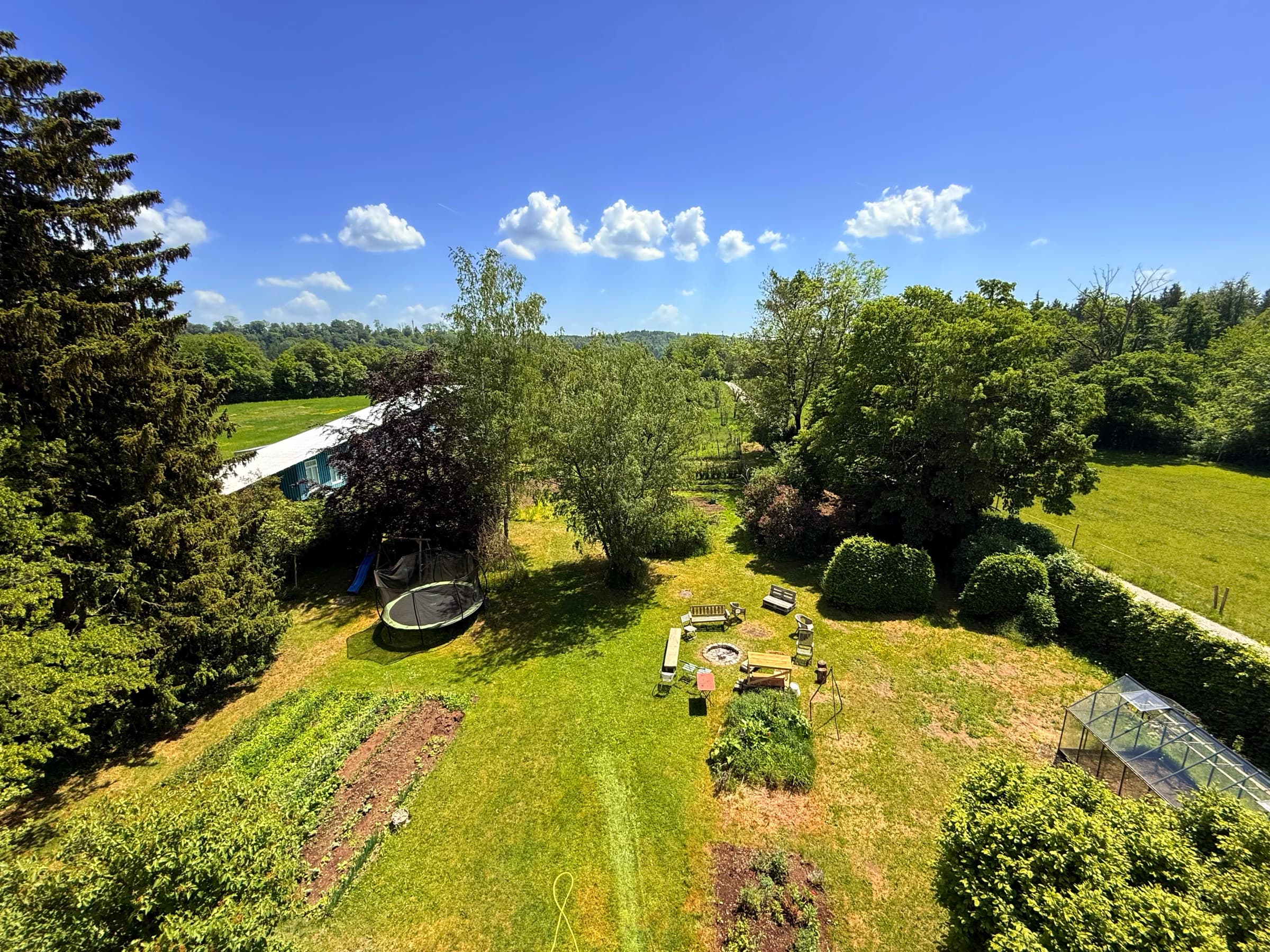 Blick über den Zehnerhof: Wiese, Garten, Trampolin, Feuerstelle, im Hintergrund Bäume und blauer Himmel mit Wolken.
